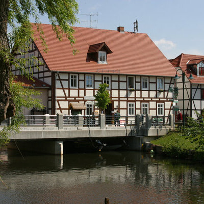 wasserseitige Ansicht auf die alte Dorfmühle. Im vorderen Bereich sehen Sie eine Brücke und dahinter das Fachwerkgebäude.