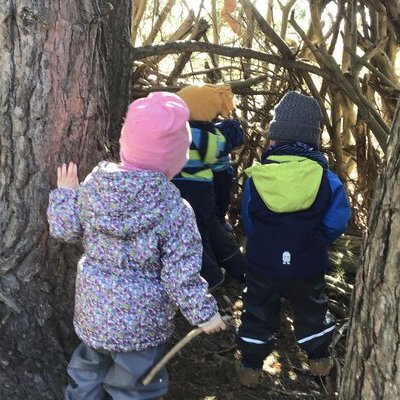 Drei Kinder gehen zwischen 2 Bäumen durch und bauen dort aus Ästen eine Bude.