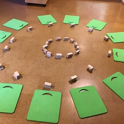 In einem Kreis liegen auf dem Boden dreizehn grüne viereckige Sitzkissen. Davor befindet sich jeweils ein Bauklotz. In der Mitte ist ein kleiner Kreis aus Bauklötzern aufgestellt.