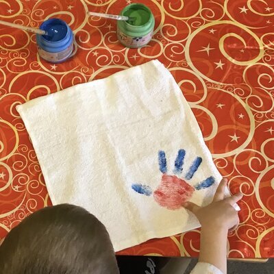 Von oben sieht man den Hinterkopf eines Kindes im linken Bildrand. Das Kind hat gerade einen blau - roten Handabdruck auf ein Stück weissen Stoff vor ihm aufgebracht.