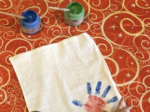 Von oben sieht man den Hinterkopf eines Kindes im linken Bildrand. Das Kind hat gerade einen blau - roten Handabdruck auf ein Stück weissen Stoff vor ihm aufgebracht.