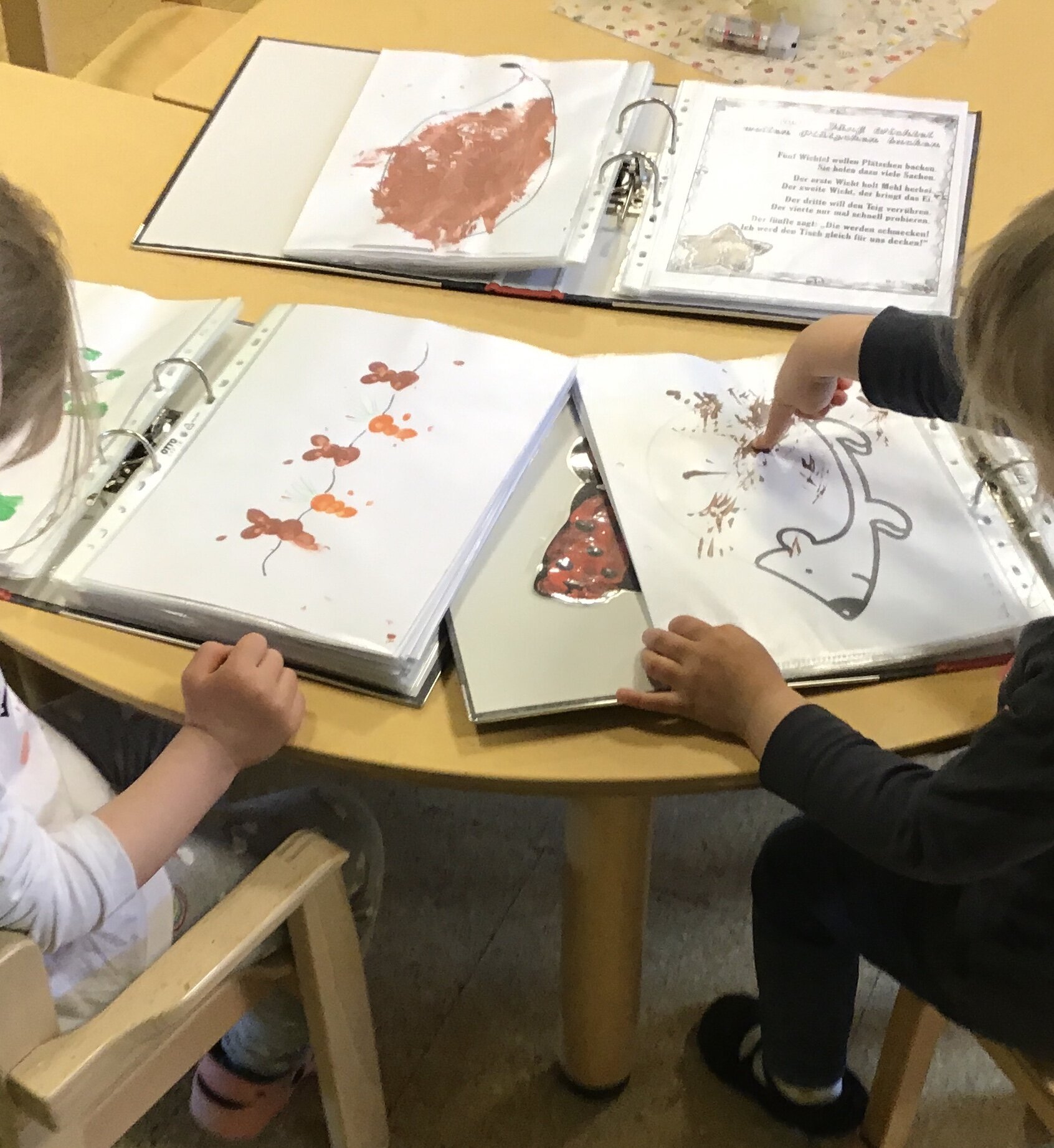 Am Seitenrand erkennt man links und rechts je ein Kind. Sie sitzen an einem Tisch´und arbeiten mit Farbe an eigenen Bildern.